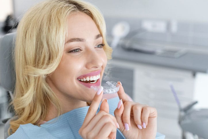 invisible braces orthodontics, modern dentistry concept. Happy young attractive blonde woman holding invisible braces or trainer while sitting at dental chair, closeup portrait